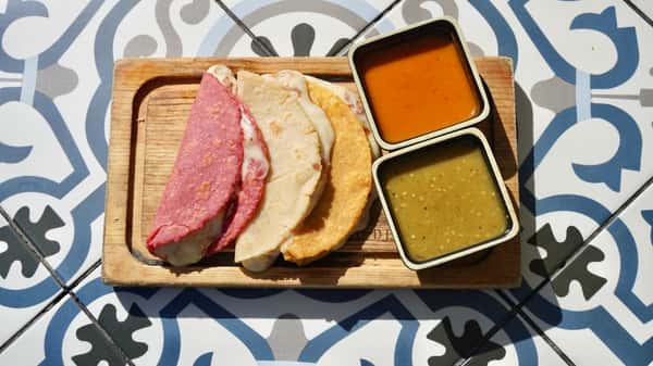 Three quesadillas made with pink, white, and yellow corn tortillas filled with melted cheese, served with red and green salsas on a rustic wooden tray.
