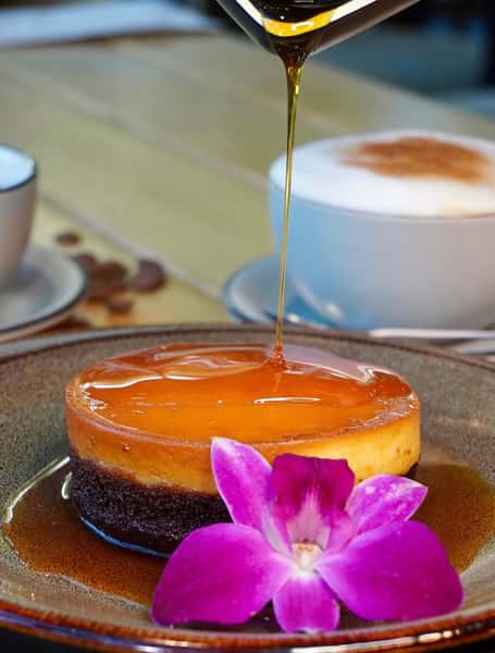 Rich chocolate and vanilla flan drizzled with caramel sauce, topped with an orchid flower, served alongside a cappuccino in a cozy café setting.