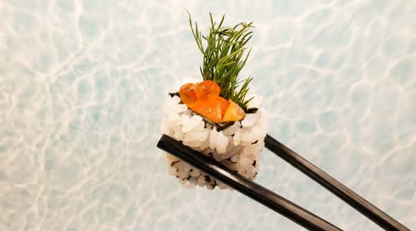 Sushi roll with white rice, orange roe, raw salmon, and green dill, held by black chopsticks.