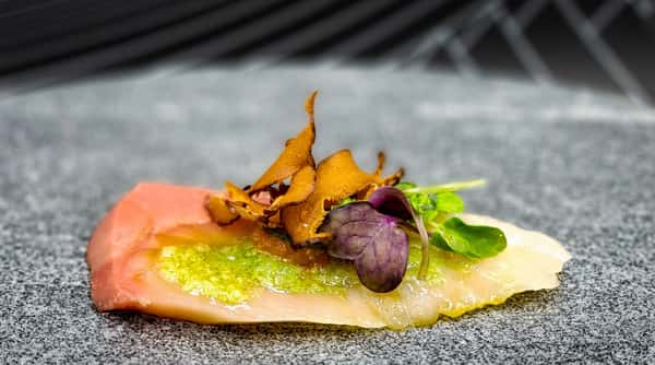 Pink and white carpaccio with crispy orange accents, green herb garnish, and a light green sauce on a grey surface.