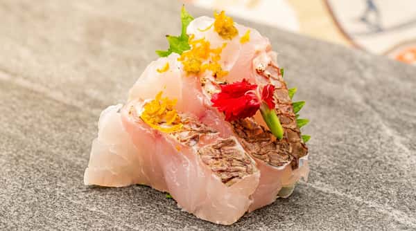 Cobia sashimi garnished with orange zest, green paste, a red flower, and green shiso leaf on a grey surface.
