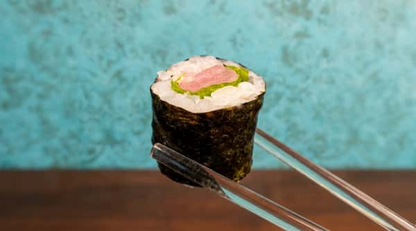 Sushi roll with white fish and green lettuce, held by black chopsticks against a turquoise background.