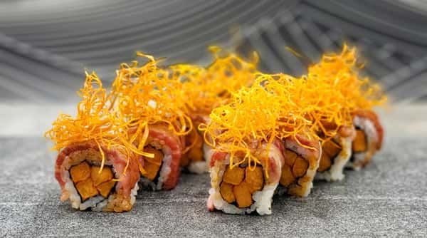 Sushi rolls wrapped in thinly sliced meat, topped with crispy orange strands, on a gray speckled surface.