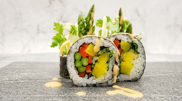 Vegetable sushi rolls with red pepper, yellow radish, peas, asparagus, and cilantro, drizzled with sauce on a grey surface.