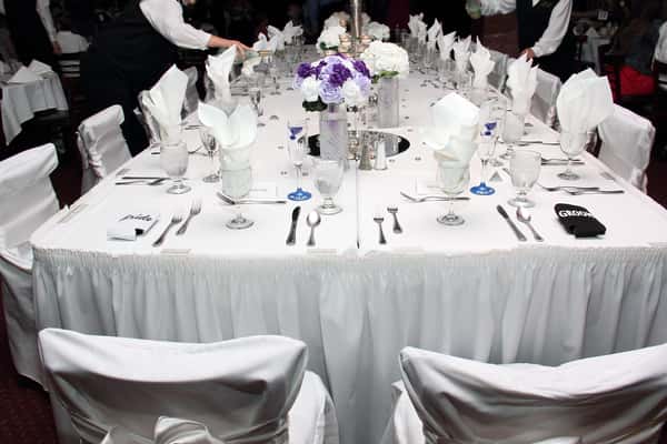 Elegant Italian‑style wedding reception room at Rose of the Hill in St. Louis, featuring long banquet tables draped in white linens, fresh rose centerpieces, greenery accents, polished chandeliers under vaulted ceilings, and historic dark wood bar in the background."