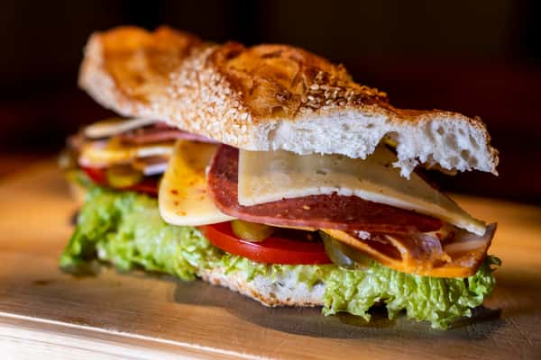 Italian sub sandwich with salami, sliced cheese, pickles, tomatoes, and lettuce on sesame-crusted bread.