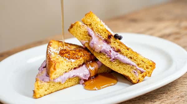 French toast sandwiches with pink filling, powdered sugar, and maple syrup pouring over on a white plate.