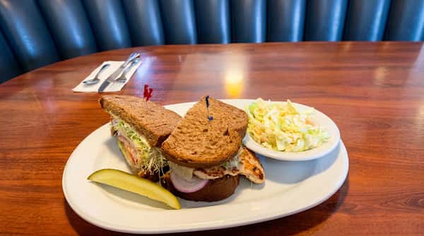 Grilled chicken sandwich on whole wheat bread with sprouts, red onion, a dill pickle spear, and coleslaw.