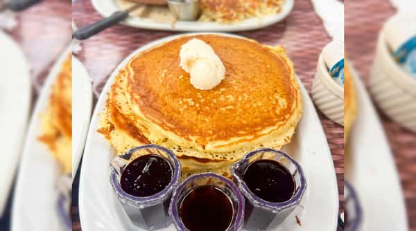 Stack of golden pancakes with a dollop of butter, served with three small pitchers of syrup/jelly.