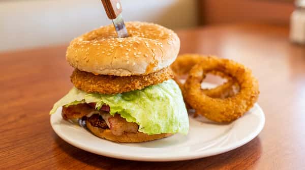 Burger with a sesame bun, fried patty, crisp lettuce, a knife spearing the bun, and onion rings on a white plate.