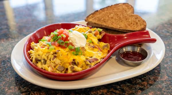 Scrambled eggs with cheese, diced tomatoes, green onions, and sour cream in a red dish, with toast and ketchup.