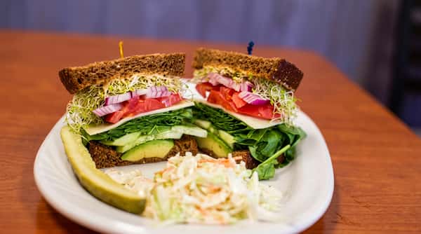 Vegetable sandwich on dark bread with sprouts, tomato, red onion, and cheese, served with coleslaw and a pickle.