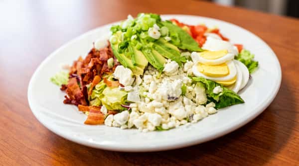 Cobb salad with bacon, avocado, crumbled cheese, sliced egg, and diced tomatoes on a white plate.
