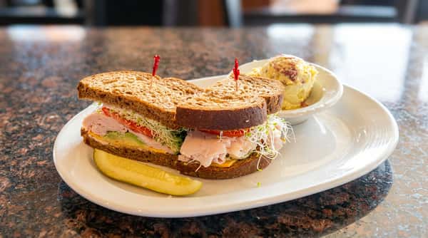 Turkey sandwich on dark bread with cheese, avocado, tomato, and sprouts, served with a pickle spear and potato salad.