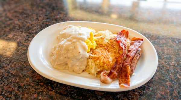 Breakfast plate with scrambled eggs, hash browns, biscuits and gravy, and strips of bacon.