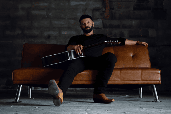 Photo of Will Dempsey sitting on a leather sofa with a guitar for his upcoming show at Smith's Olde Bar on October 8, 2026