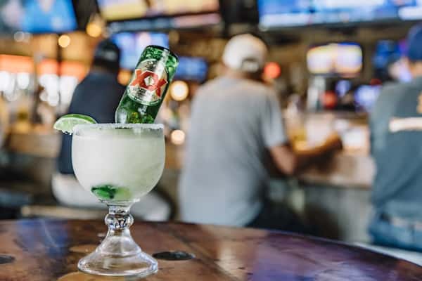 Frozen margarita with upside-down Dos Equis bottle and lime wedge on bar, sports fans blurred in background