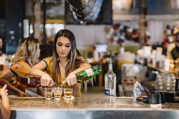 Bartender pouring liquor into three shot glasses behind a busy bar counter with bottles and patrons in background.