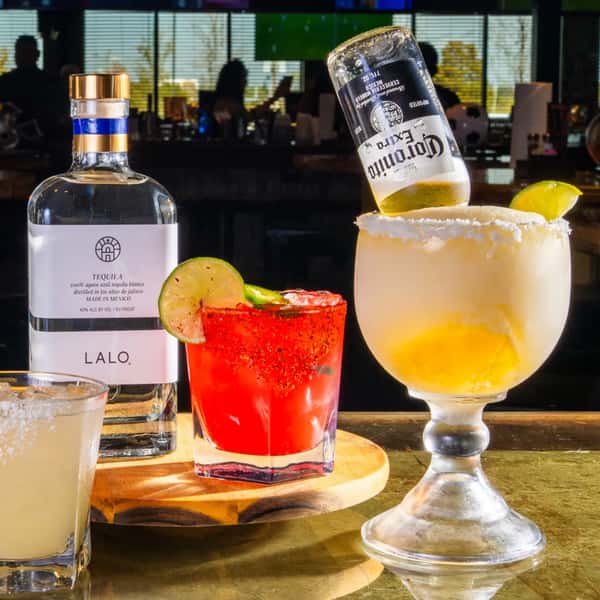 LALO tequila bottle with red cocktail and Corona-topped margarita, lime wedges and salt rims on bar counter