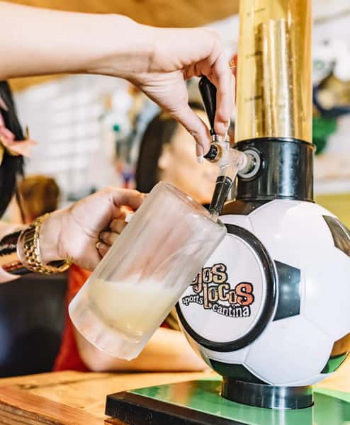 Hand pours draft beer into frosty mug from OjOs Sports Cantina soccer-themed tap tower