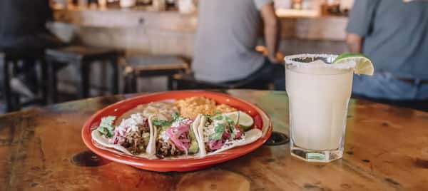 Three street tacos with cilantro and pickled onions on a red plate beside a salt-rimmed margarita with lime wedge