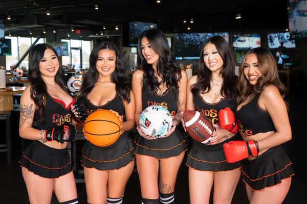Five women in OjOs Locos uniforms pose with basketball, soccer ball, football, and boxing gloves inside sports bar