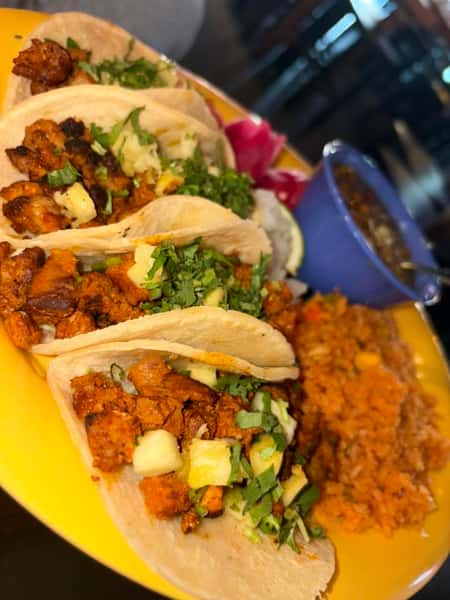 Four street tacos with seasoned pork, pineapple, and cilantro, served with rice and beans on a yellow plate