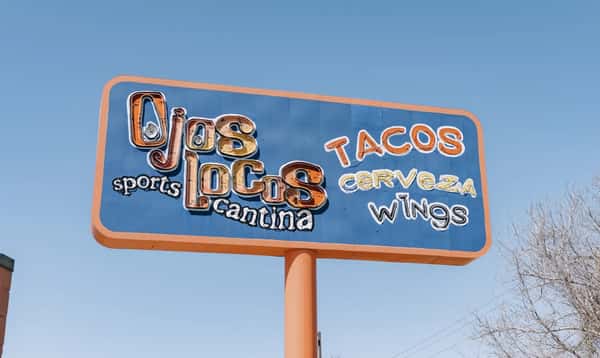 OjOS Locos Sports Cantina roadside sign advertising tacos, cerveza, and wings against a clear blue sky