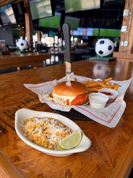 Crispy chicken sandwich with fries and dipping sauces, knife through bun, esquites with lime on wooden table in sports bar