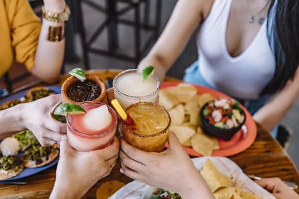 Friends clink colorful margaritas with lime wedges over tacos and chips at a lively Mexican restaurant table