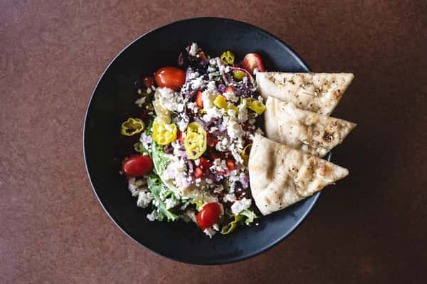 Greek salad with cherry tomatoes, olives, feta cheese, banana peppers, and flatbread slices.