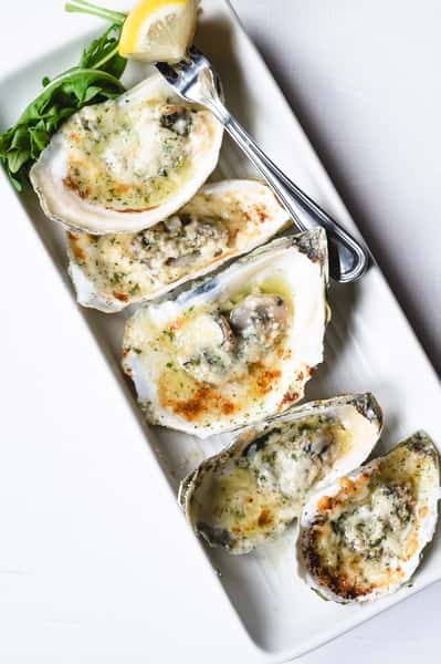 Baked oysters in their shells, topped with melted cheese and herbs, served on a rectangular white platter with a lemon wedge and greens.