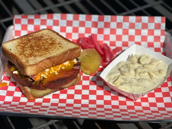 Grilled bologna sandwich with melted cheese, pickles, and sauce on toasted bread served with creamy pasta salad, pickled onions, and pickle slices on a checkered tray