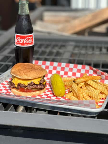 Classic cheeseburger with crinkle fries and pickle served with a glass bottle Coca-Cola on a checkered tray