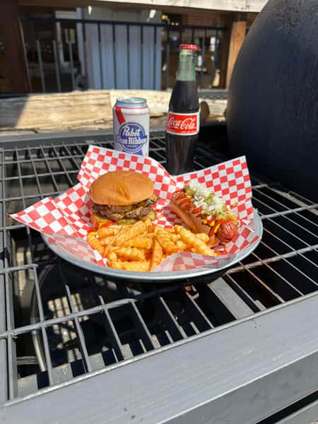 Classic cheeseburger, hot dog, and crinkle fries with soda and beer served outdoors at a casual American barbecue restaurant
