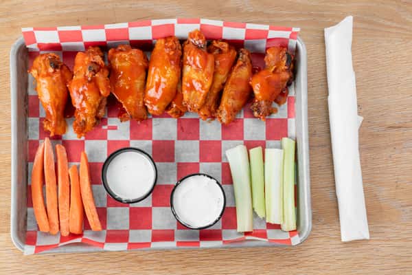Buffalo-style chicken wings with a red-orange glaze served on red and white checkered paper in a metal tray with dipping sauce cups and celery sticks at a restaurant