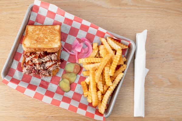 The Pig Melt sandwich served in a metal tray with crinkle-cut french fries, pickled pink onions, and pickle slices on red and white checkered paper at a restaurant