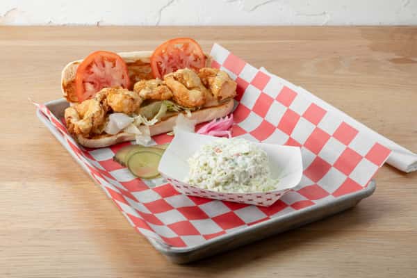 Fried shrimp served on red and white checkered paper in a metal tray with a side of coleslaw, pickles, and tomato slices on a wooden surface at a restaurant