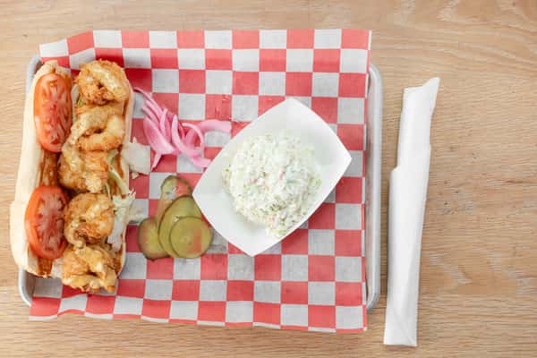 Fried shrimp po'boy served on red and white checkered paper with a small bowl of coleslaw, pickled pink onions, pickle slices, and a wrapped utensil at a restaurant