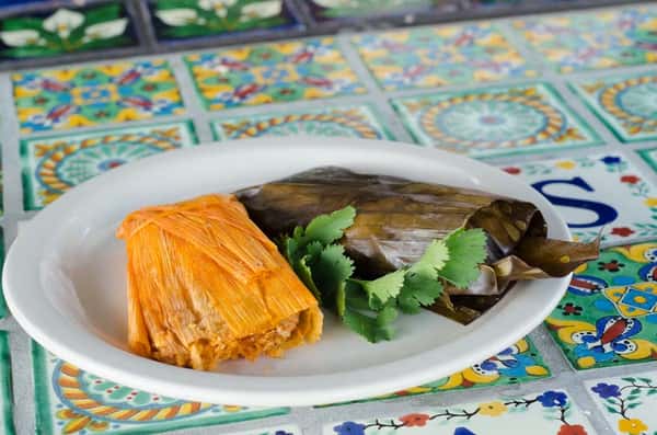 Traditional Mexican tamales wrapped in corn husk and banana leaf, served on a white plate with fresh cilantro – authentic homemade Mexican cuisine on a colorful tiled table.