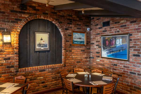 Warm, nautical-themed dining nook at Sindbad's Restaurant, featuring exposed brick walls, maritime artwork including lighthouse and boat paintings, and a round wooden table set for six — creating a cozy, seafaring ambiance.