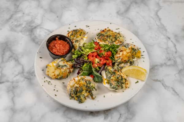 Top-down view of Oysters Rockefeller baked with spinach and cheese, served with a lemon wedge, fresh greens, diced tomatoes, and cocktail sauce — a savory seafood appetizer.