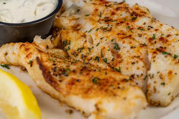 Close-up of seasoned grilled white fish fillets garnished with herbs, served with a wedge of lemon and creamy tartar sauce — a light and flavorful seafood entrée.