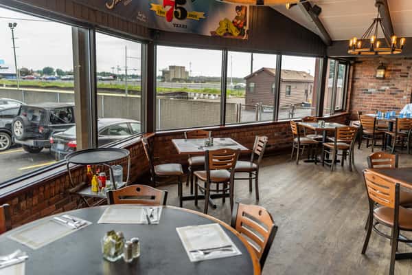 Cozy casual dining area with rustic brick walls, large panoramic windows offering a view of the parking lot and highway, and wooden tables set with placemats and condiments — ideal for a relaxed family meal or classic American-Italian dining experience.