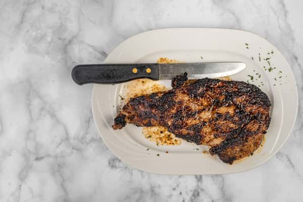 Grilled blackened ribeye steak with a seared crust served on a white plate, accompanied by a steak knife — a bold and flavorful cut of beef.