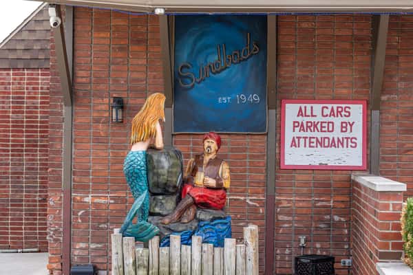 Decorative outdoor statue at Sindbad's Restaurant entrance, featuring a mermaid and sailor sculpture below a sign reading "Sindbad's Est. 1949" — a whimsical nod to nautical folklore.