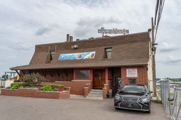 Exterior of Sindbad's Restaurant, a long-standing Detroit waterfront dining spot, featuring a brown shingle roof, red brick facade, and neon signage, with valet parking and landscaped greenery at the entrance.