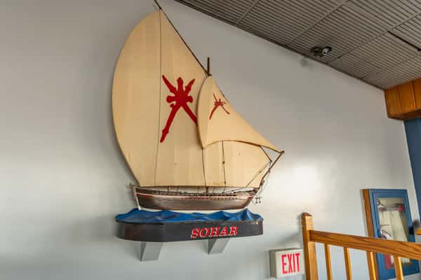 Wall-mounted model of the traditional Omani sailing ship "Sohar" with tan sails featuring red insignias, displayed above an EXIT sign inside Sindbad's Restaurant — a nautical-themed decor element celebrating maritime heritage.