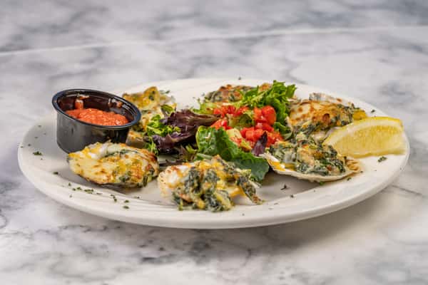 Full plate of baked Oysters Rockefeller with melted cheese and spinach, served with mixed greens, diced tomatoes, a lemon wedge, and cocktail sauce.