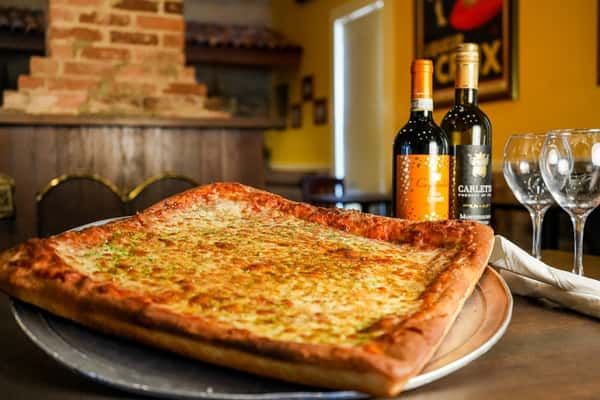 Large rectangular cheese pizza on a tray with wine bottles and glasses in a cozy Italian restaurant setting.